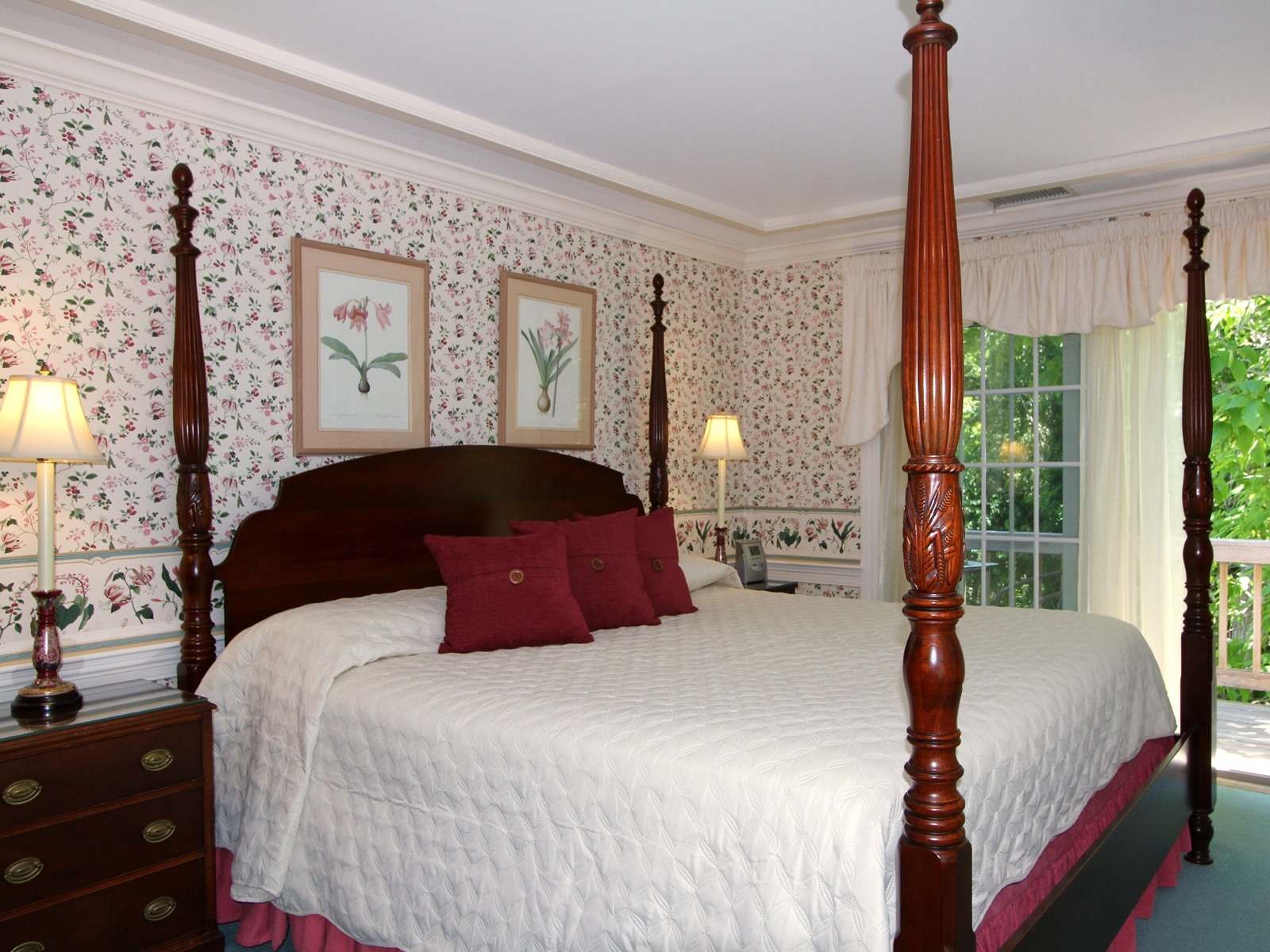 Four poster King Bed with dusty pink dust ruffle and burgundy velvet decorative pillows, view of balcony, bedside storage and candlestick lamps with white shades, green carpet and white wallpaper with pink lilies and
