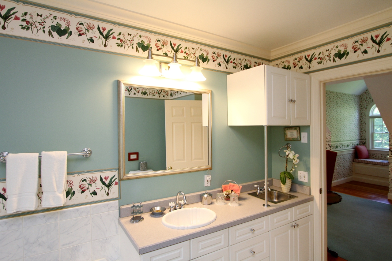 close up of vanity with white ceramice sink and stainless steel wet bar sink, fridge tucked away in cabinet above the smaller sink, decorative mirror and 3 vanity light fixture above main sink