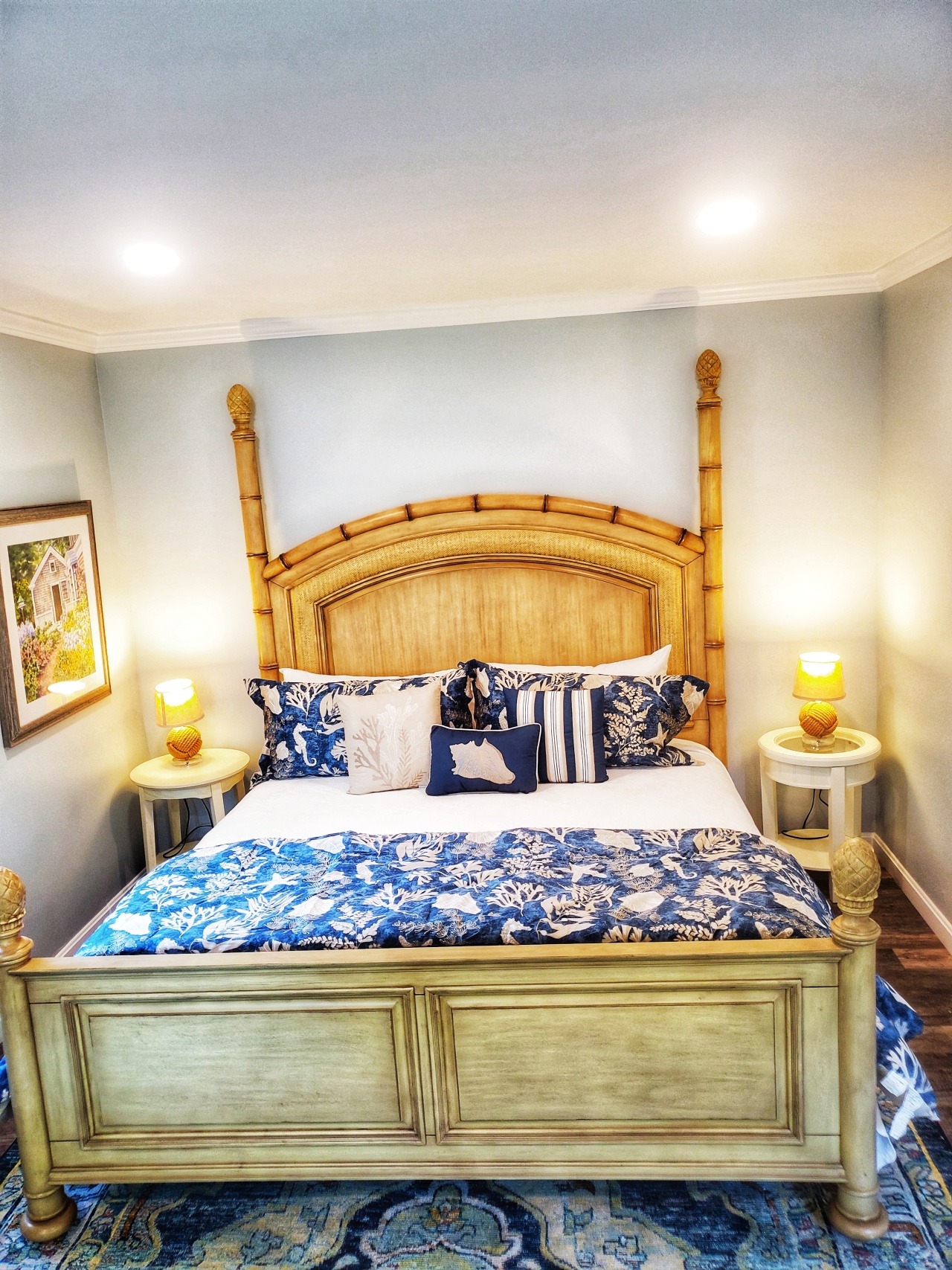 Pale beachy Tommy Bahama brand King size bed with navy blue and shells bedding and decorative pillows, round white side tables with rope wrapped lamps, blue and yellow coordinating rug, pale blue grey walls and 2 recessed lights above