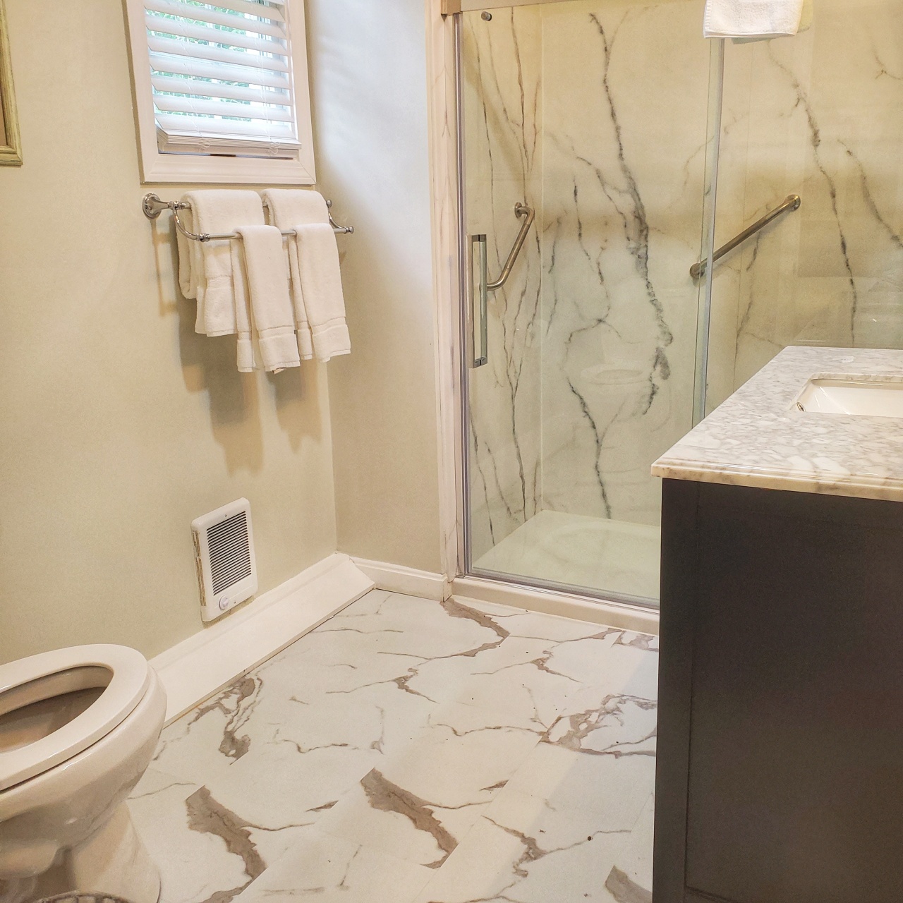 view of bathroom with marble pattern flooring, white toilet, window with blinds, and towel rack with white towels, oversize shower with grab rails
