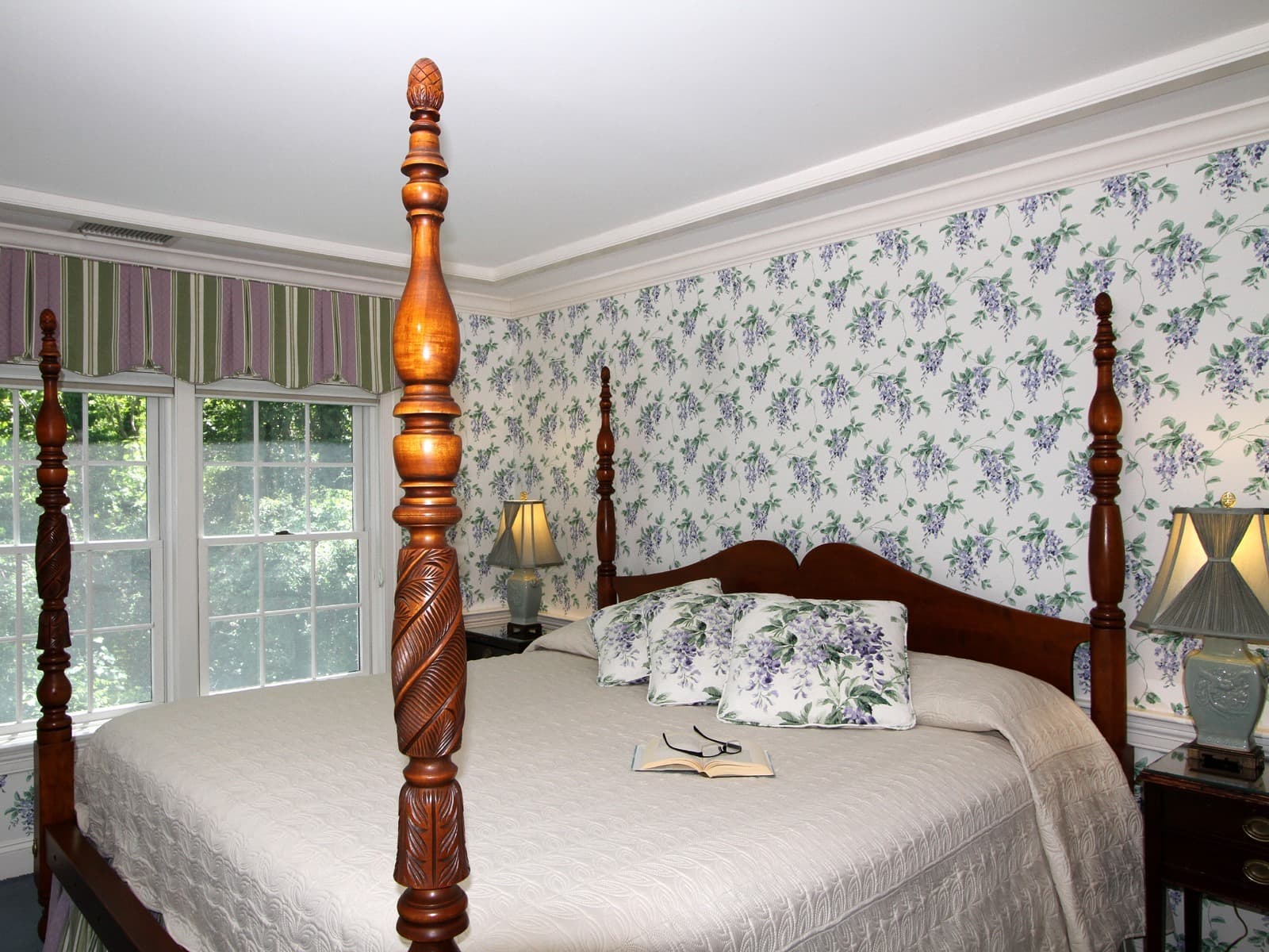 Four poster King bed with green bedding and decorative pillows, wooden side tables with lamps, white wallpaper with purple florals