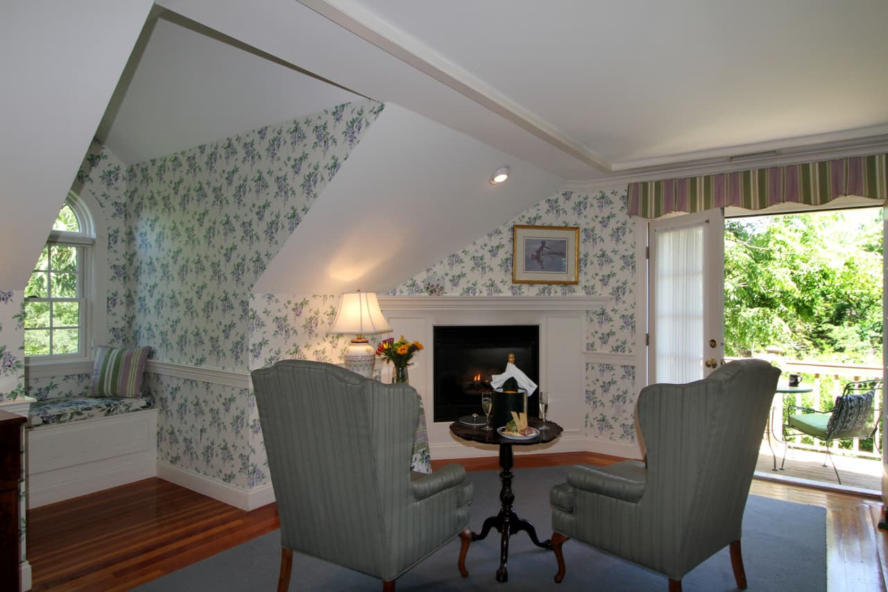 Sitting area in front of the gas fireplace with green wingback chairs, window seat with decorative pillows on bench, wooden floors with green carpeting, white with purple and green wisteria flowers wallpaper