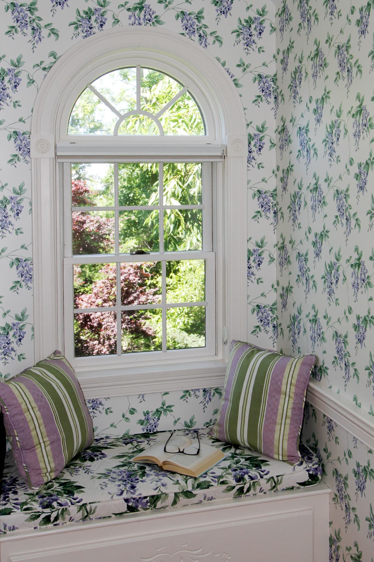 up close view of window seat bench with pillows and view out the large window of greenery and blooming flowers