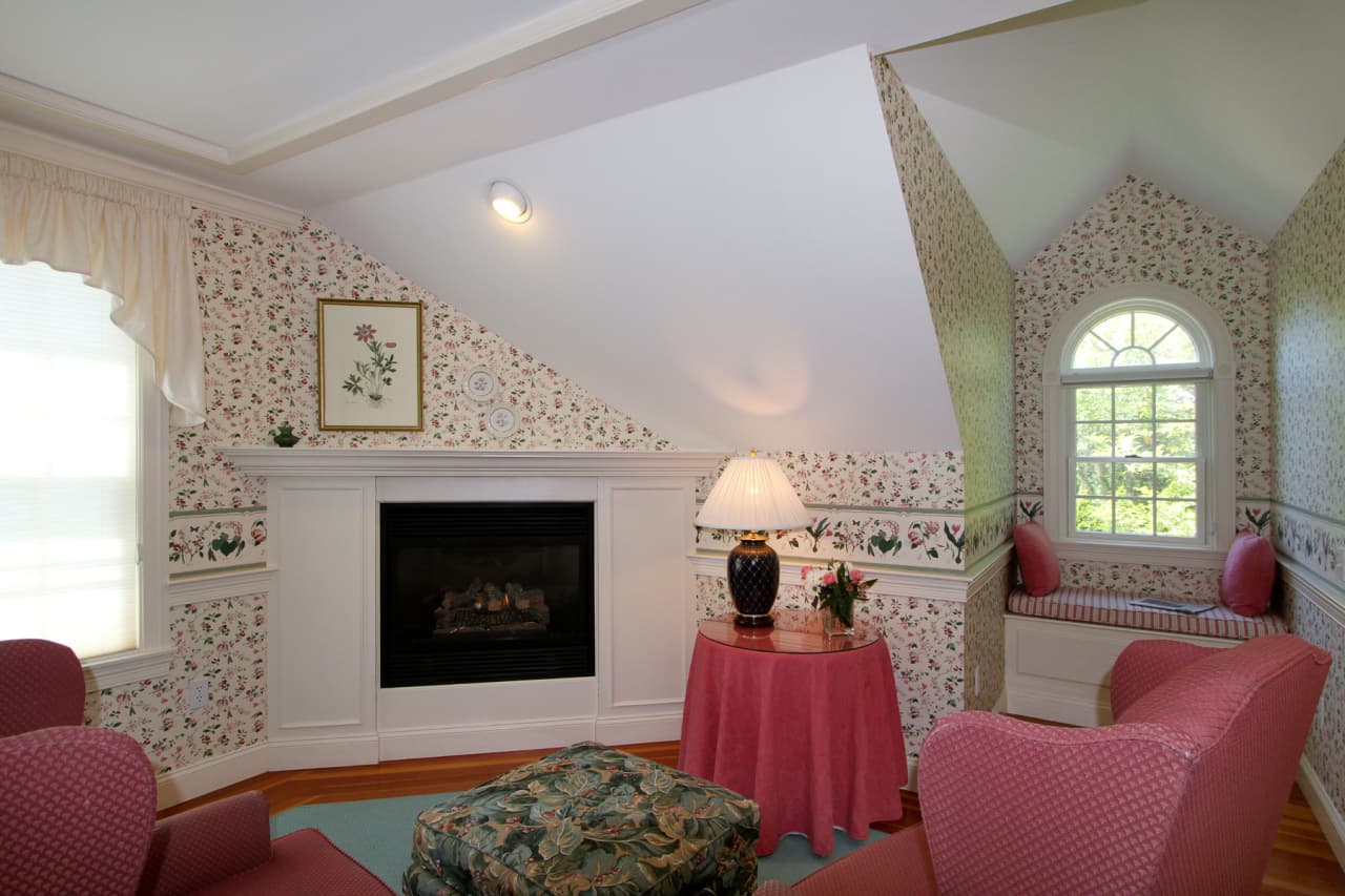 close up of sitting area with gas fireplace, burgundy wingback chairs, ottoman, table with burgundy tablecloth and burgundy lamp with white shade, view of window seat with decorative pillows, and large window