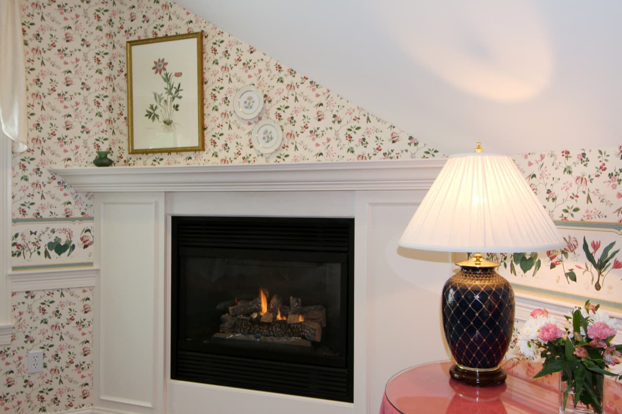 close up of fireplace and white wood mantle, table with burgundy lamp and fresh floral bouqet, framed illustration of lily above the mantle