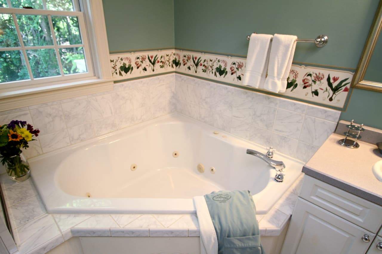 close up of white corner spa tub with white ceramic surrounding, jets and faucet in view floral wallpaper border, and muted green walls, towel bar with 2 white towels