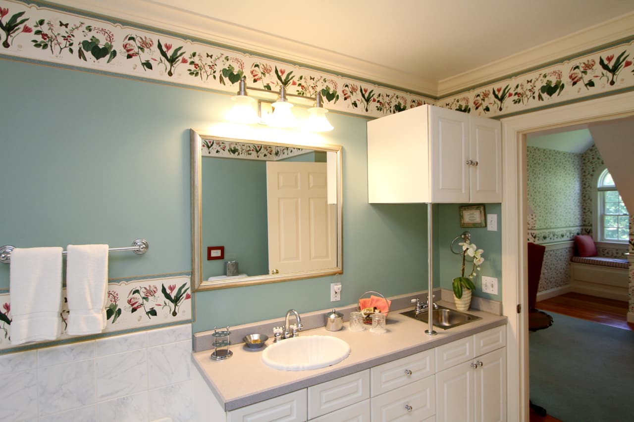 close up of vanity with white ceramice sink and stainless steel wet bar sink, fridge tucked away in cabinet above the smaller sink, decorative mirror and 3 vanity light fixture above main sink