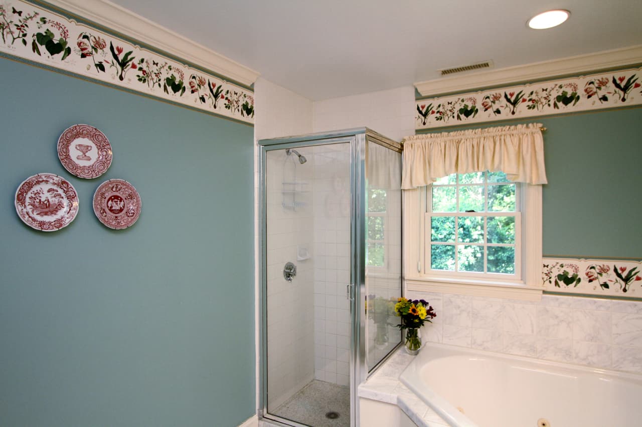 close up of shower stall with enclosed glass, window with shades and white valance, mute green walls, with floral wallpaper border, and ceramic plates in shades of burgundy decoration on the wall