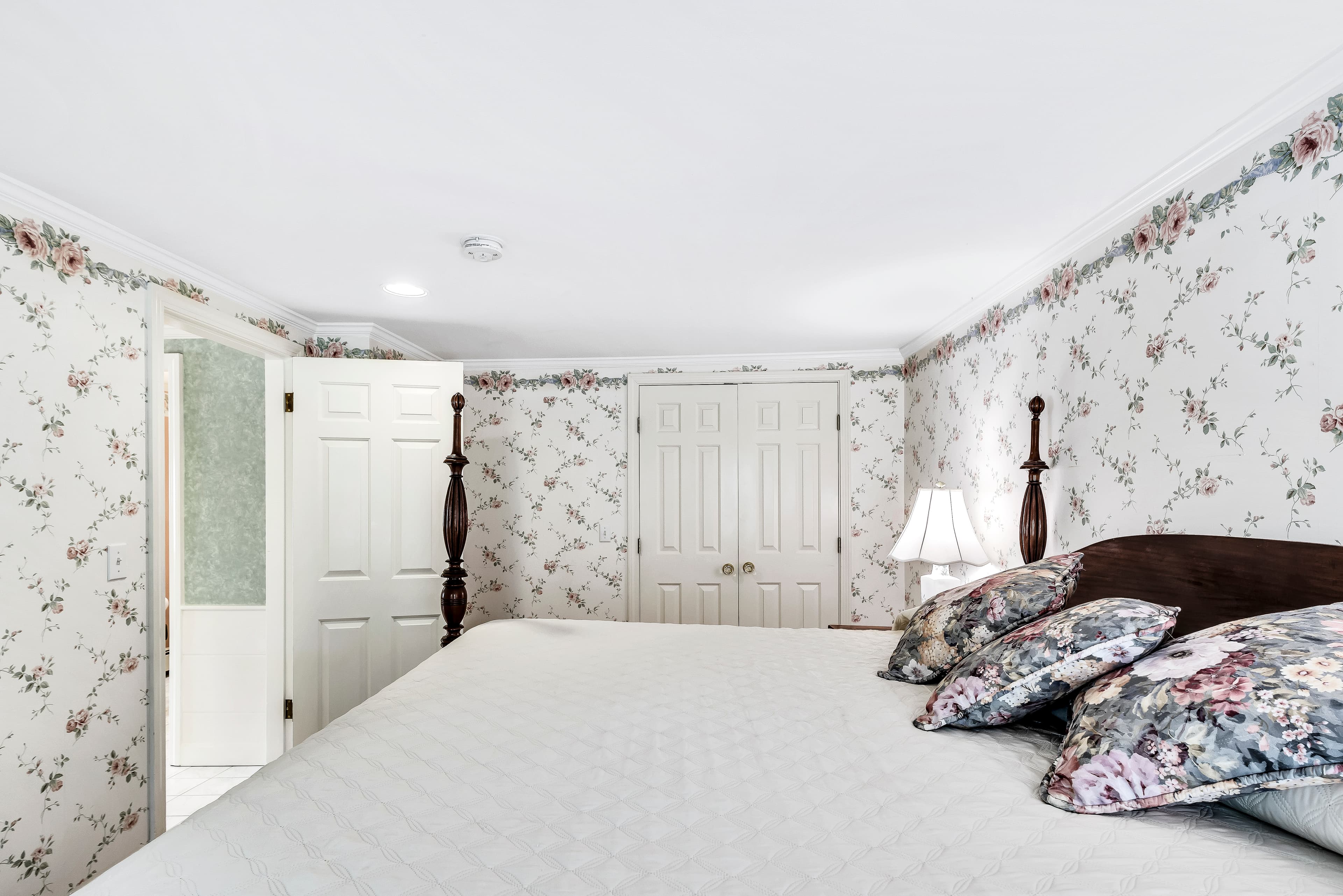 Living room area with 2 tables and table lamps, 3 wingback chairs that coordinate with the decor and decorative pillows, wooden coffee table, large window with blinds and valences, light green wallpaper and rug over hardwood floors