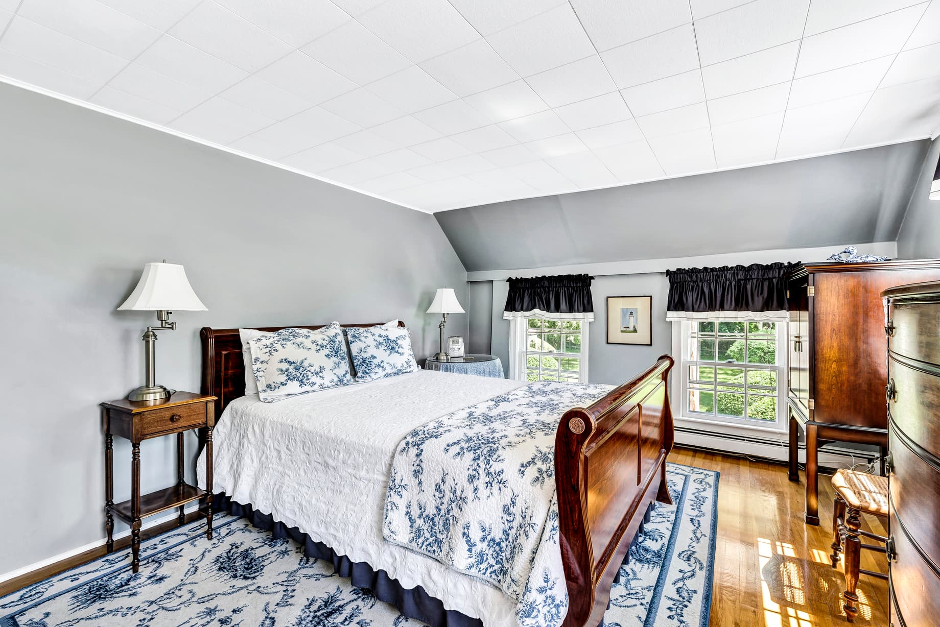 Picture of wall that separates bedroom are and living room area as well as bed and bedside lamp, window with navy valance, white sofa with navy pillows, fireplace, white and blue decorative ceramics, standing lamp