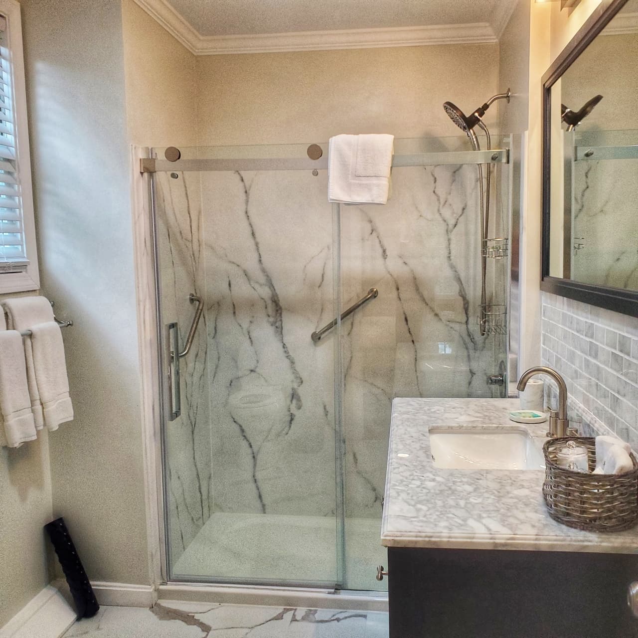 close up of oversize shower with marble decor walls and sliding glass door,grab rails, vanity with marble and marble backsplash, matching mirror above, towel rack with white towels