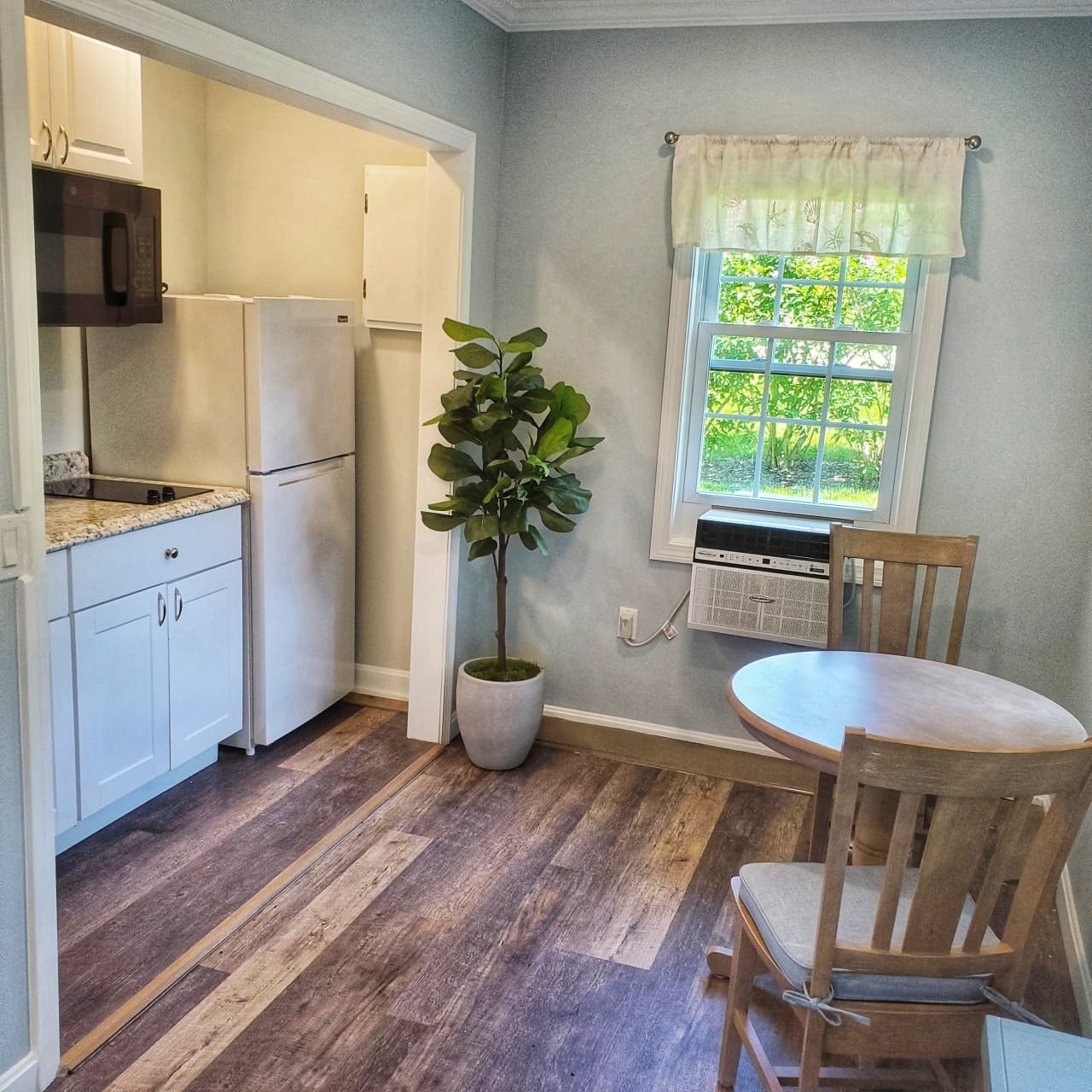 view of kitchen with counter, cook top, microwave, cabinets, fullsize refirgerator, plant and planter, window with low profile cooling unit, window valance, sitting area with light grey table for two with matching chairs