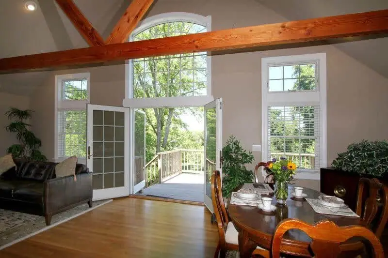 view of living room/ dining room area with large French doors to balcony area open, wood floors, wooden beams and more windows next to the balcony door shown as well