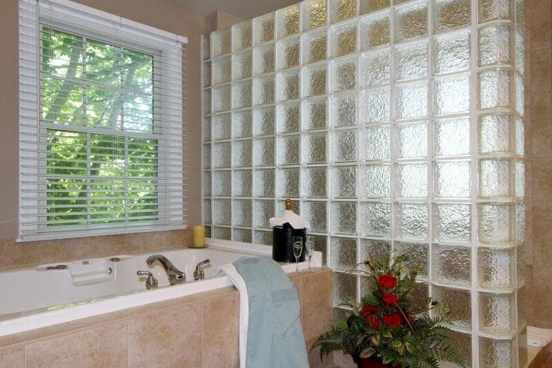 view of white spa tub, glass tiles separating the large shower area, window with blinds, beige ceramic tiling shown