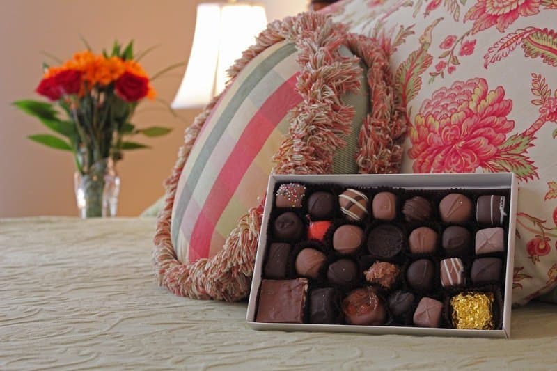 close up of decorative pillows arranged on the bed with large box of local chocolates and floral arrangement