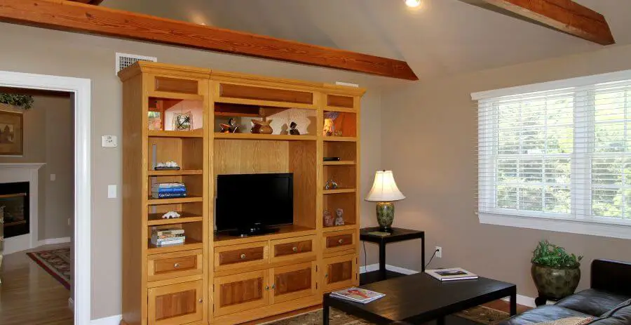 view of the living room area light wood credenza with focus lighting and flat screen TV, decorative objects on shelves and books, large window with white blinds and exposed wood beams, dark wood side table and matching coffee, table lamp