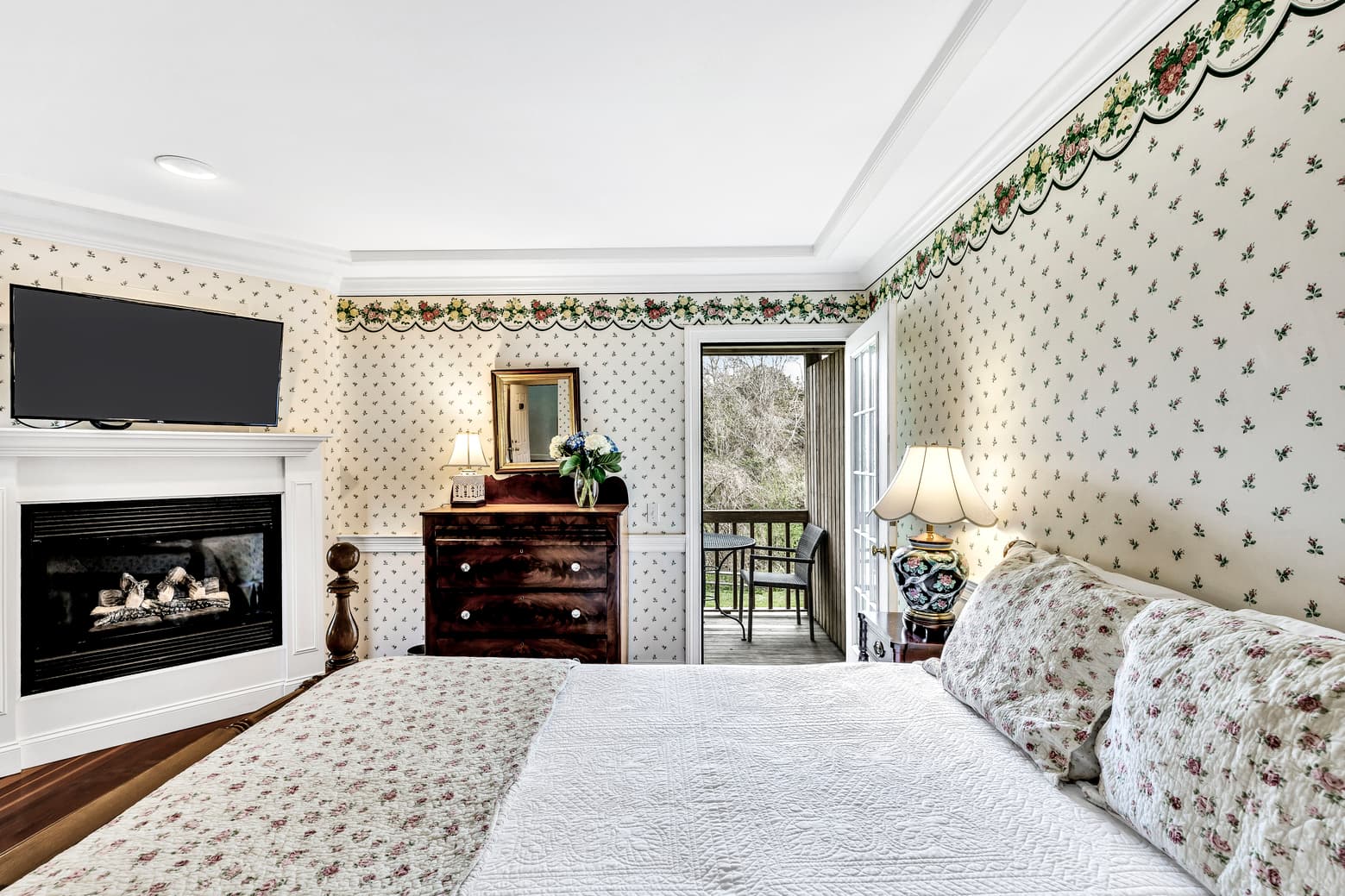 Queen bed with white and small pink roses accent bedding, door to balcony with view, dresser with mirror above, fireplace, flatscreen TV, white with small pink roses wallpaper patter