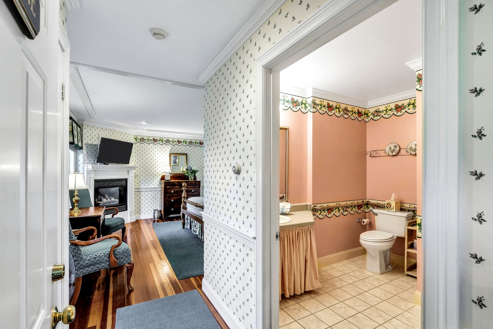 Queen bed with white and small pink roses accent bedding, door to balcony with view, dresser with mirror above, fireplace, flatscreen TV, sitting area with table and lamp,white with small pink roses wallpaper pattern