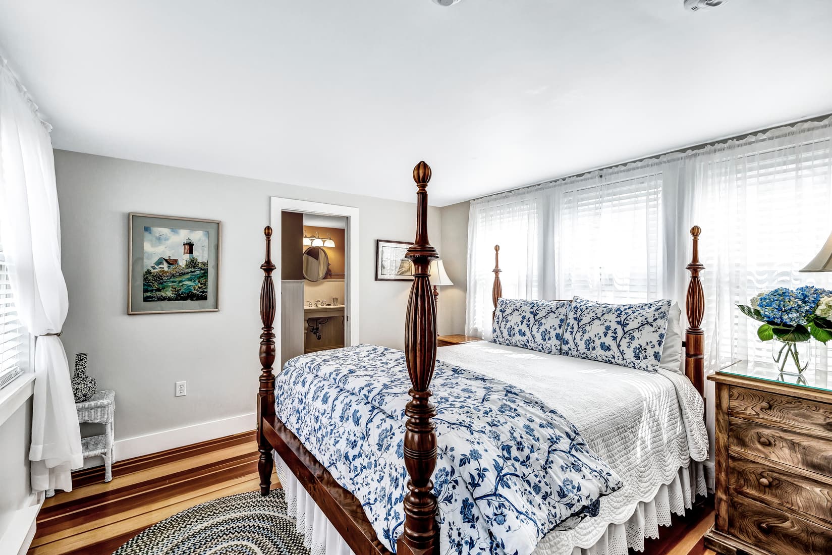 A light-filled bedroom with a four-poster bed. The bed has a white comforter with a blue and white patterned blanket and pillows.