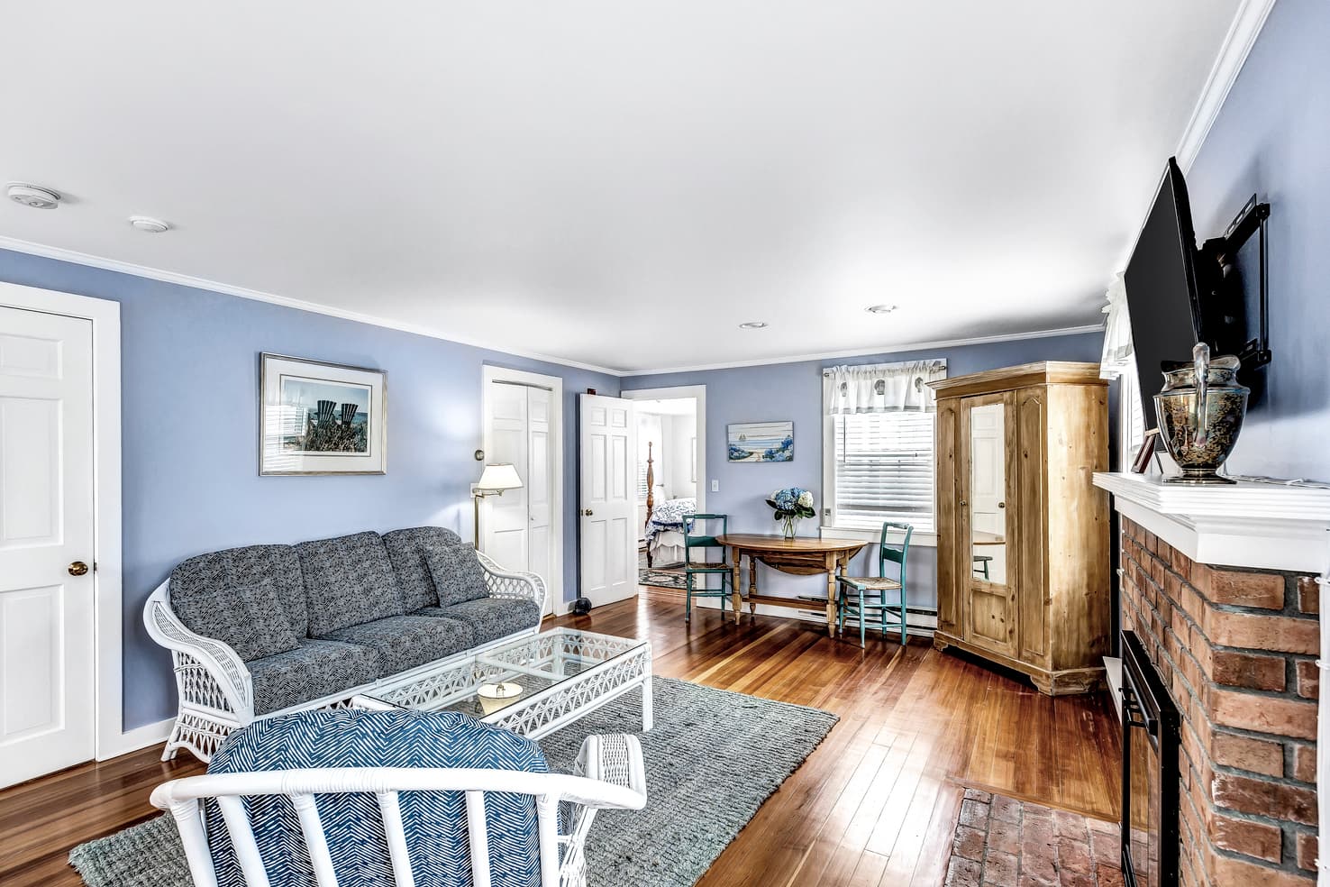A cozy living room with light blue walls, hardwood floors, and a stone fireplace. The room is furnished with a white wicker sofa and chairs.