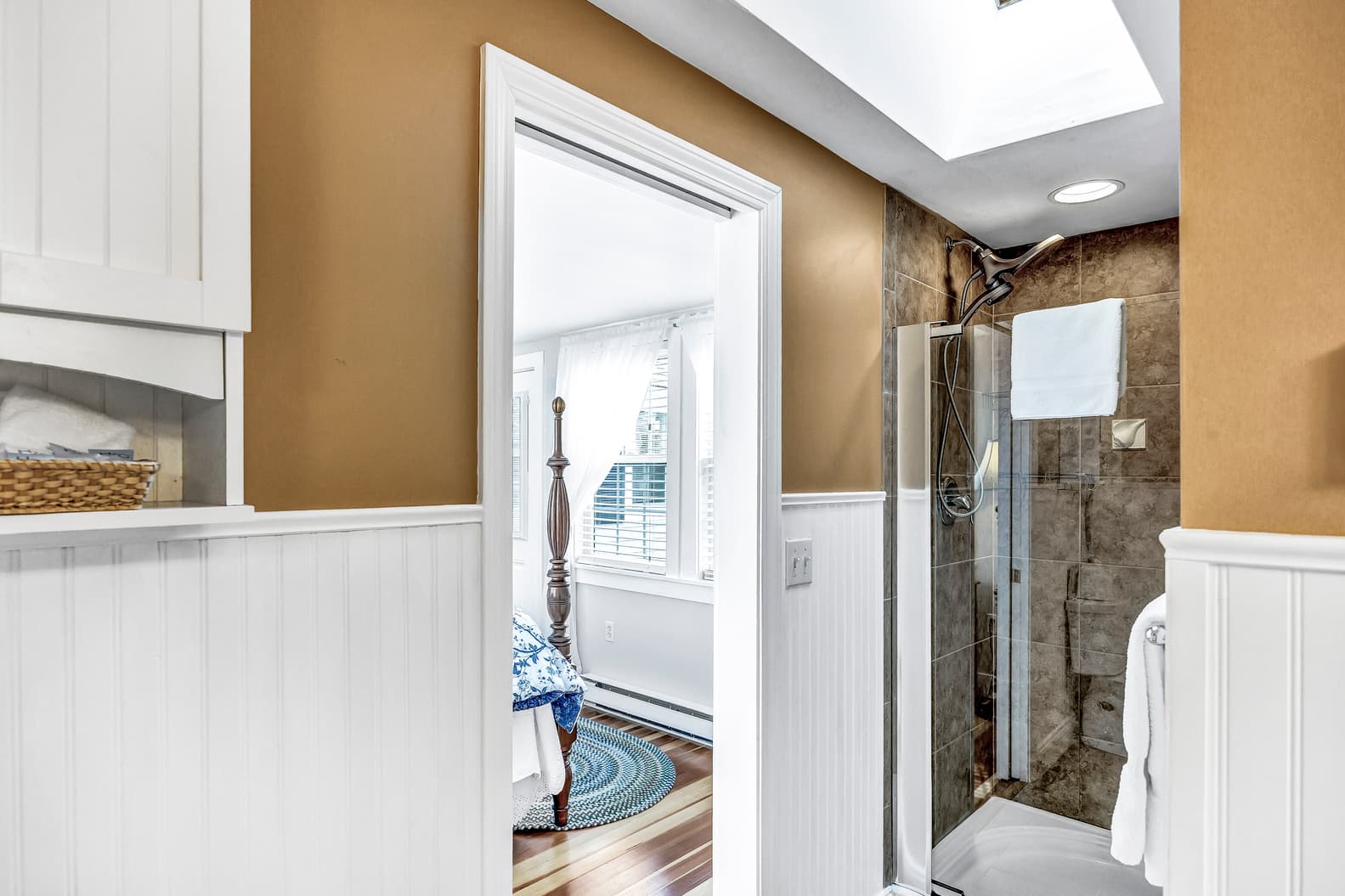 A brown and white bathroom with a glass shower stall and a view into a bedroom with a four-poster bed.
