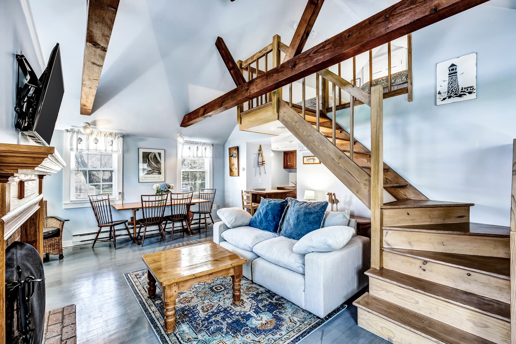 Living room/ dining room area with gray stained wooden floors, white down filled sofa with oversize denim pillows, wooden square coffee table, wooden full size dining table and 4 chairs, stairs leading to loft area, light blue painted walls with 2 windows decorated with white blinds shown
