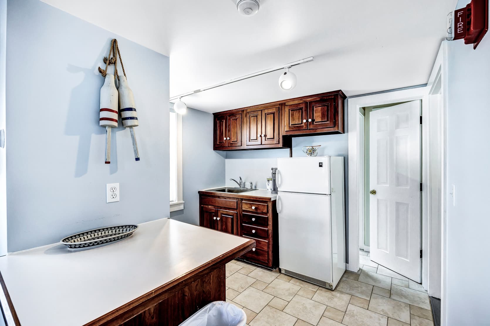 Full size kitchen with dark wood cabinets, stainless steel sink, Kcup coffee machine, fullsize white fridge, ceramic tile floor, door to bathroom area, white round lighting fixtures