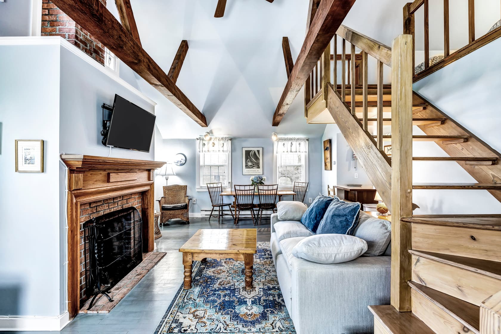Living room area with focus on wood burning fireplace with wooden mantle, flatscreen TV above, exposed wood beams for decor, windows with white blinds and window treatments, Mistubushi ductless A/C and heating unit