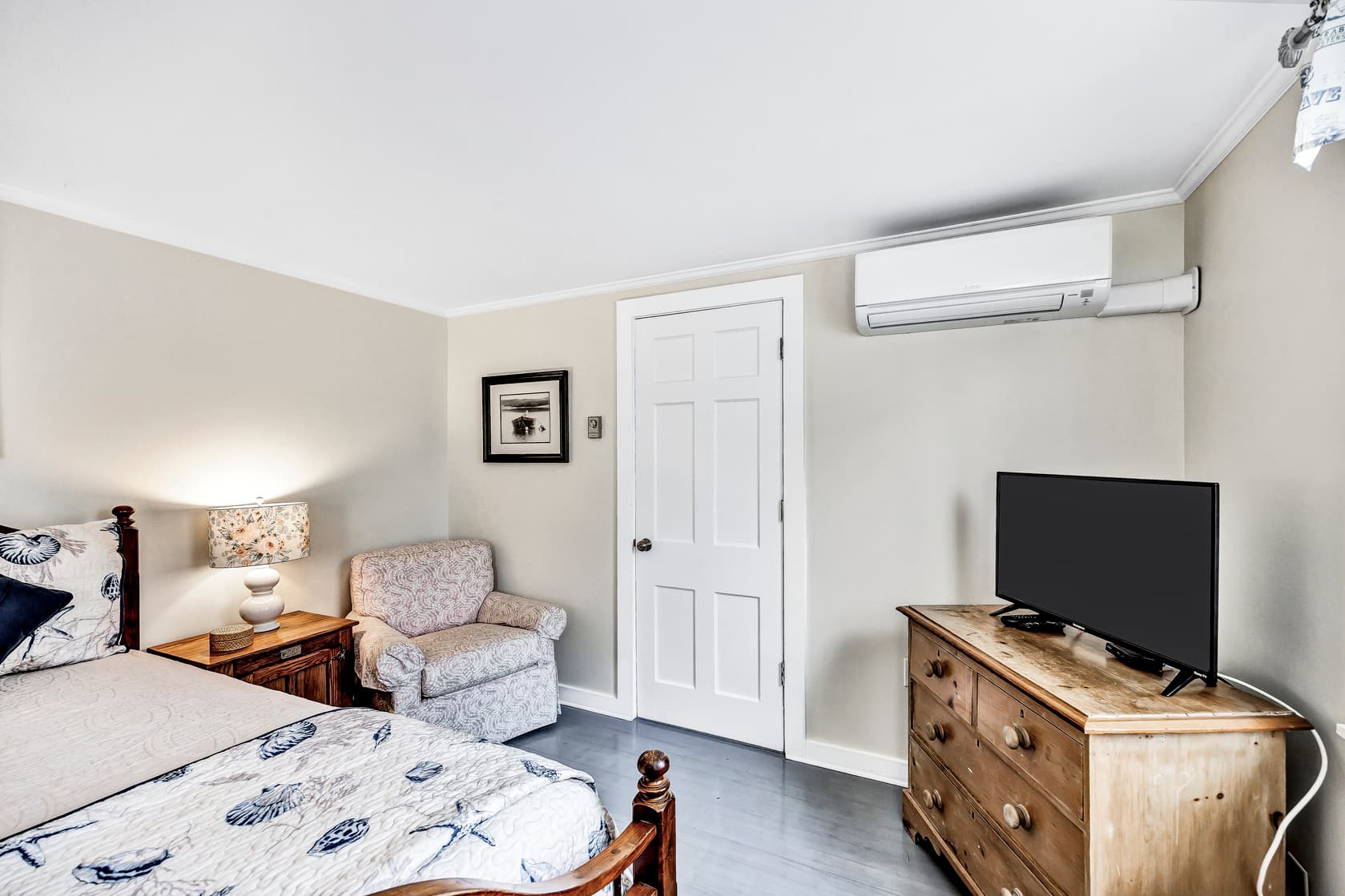 queen bed with coastal decor pillows and bed side table and lamp, reading chair,dreser, flatscreen TV, ductless A/C