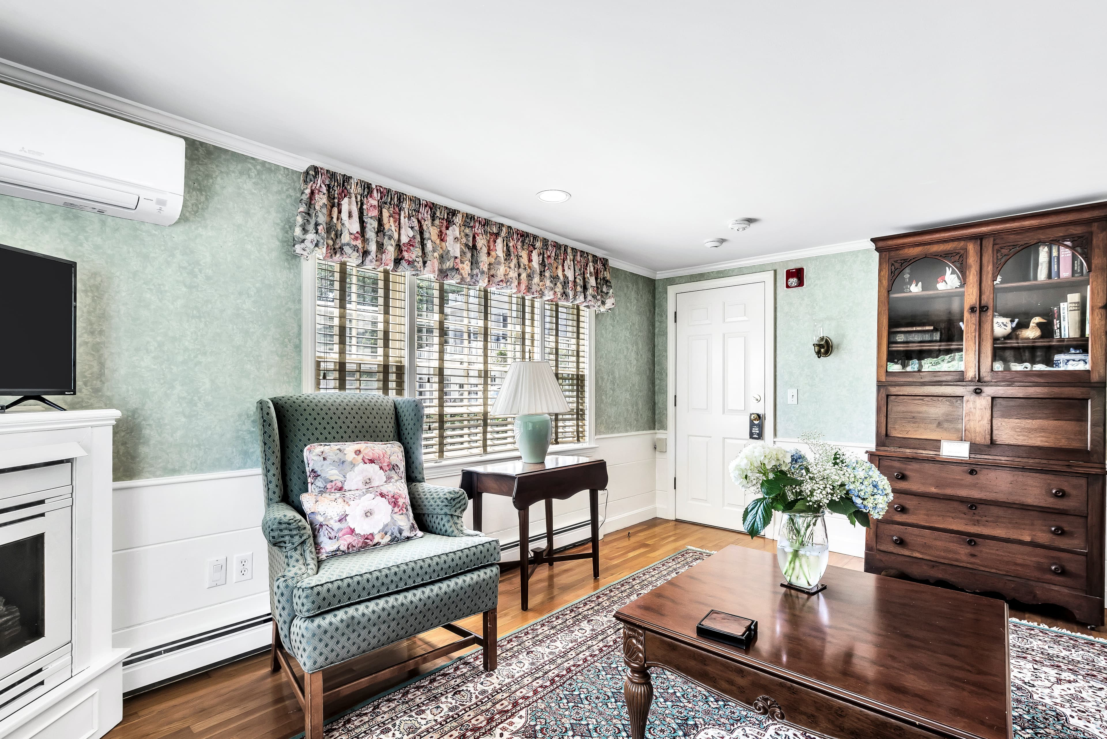 close up of sitting area with wingback chairs, table and lamp, tablecloth and decorative pillows match and coordinate with light green wallpaper and chair rail moldings, picture of saltbox cottage with florals in bloom above the chairs on the wall