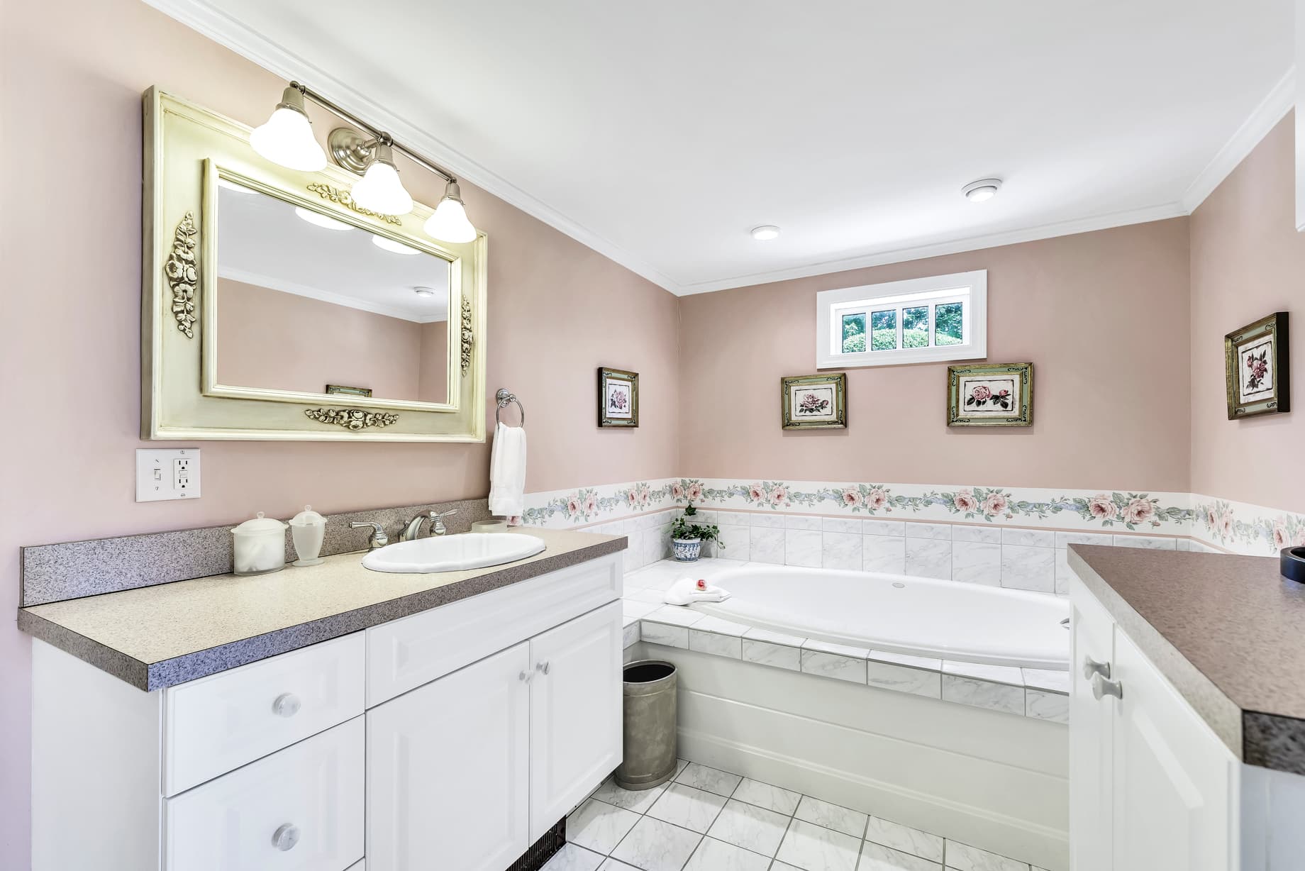 close up of 4 poster King bed with floral dust ruffle and matching decorative pillows, wooden side tables with lamps, and white with little pink roses wallpaper, window with blinds
