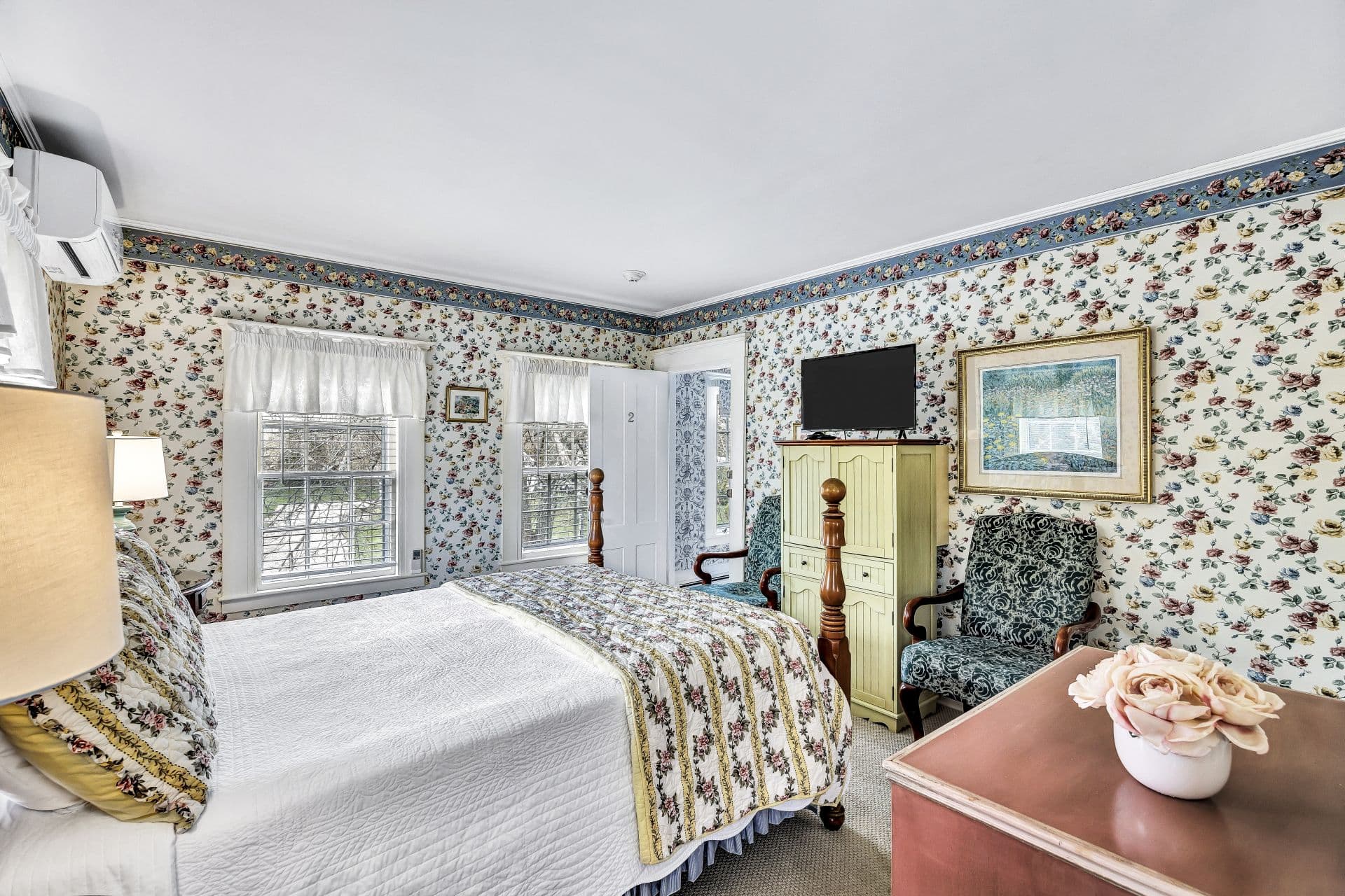 side view of Queen 4 poster bed with yellow stripes and roses bedding, white with yellow, pink and blue roses wall paper, dresser, green sitting chair and flatscreen TV