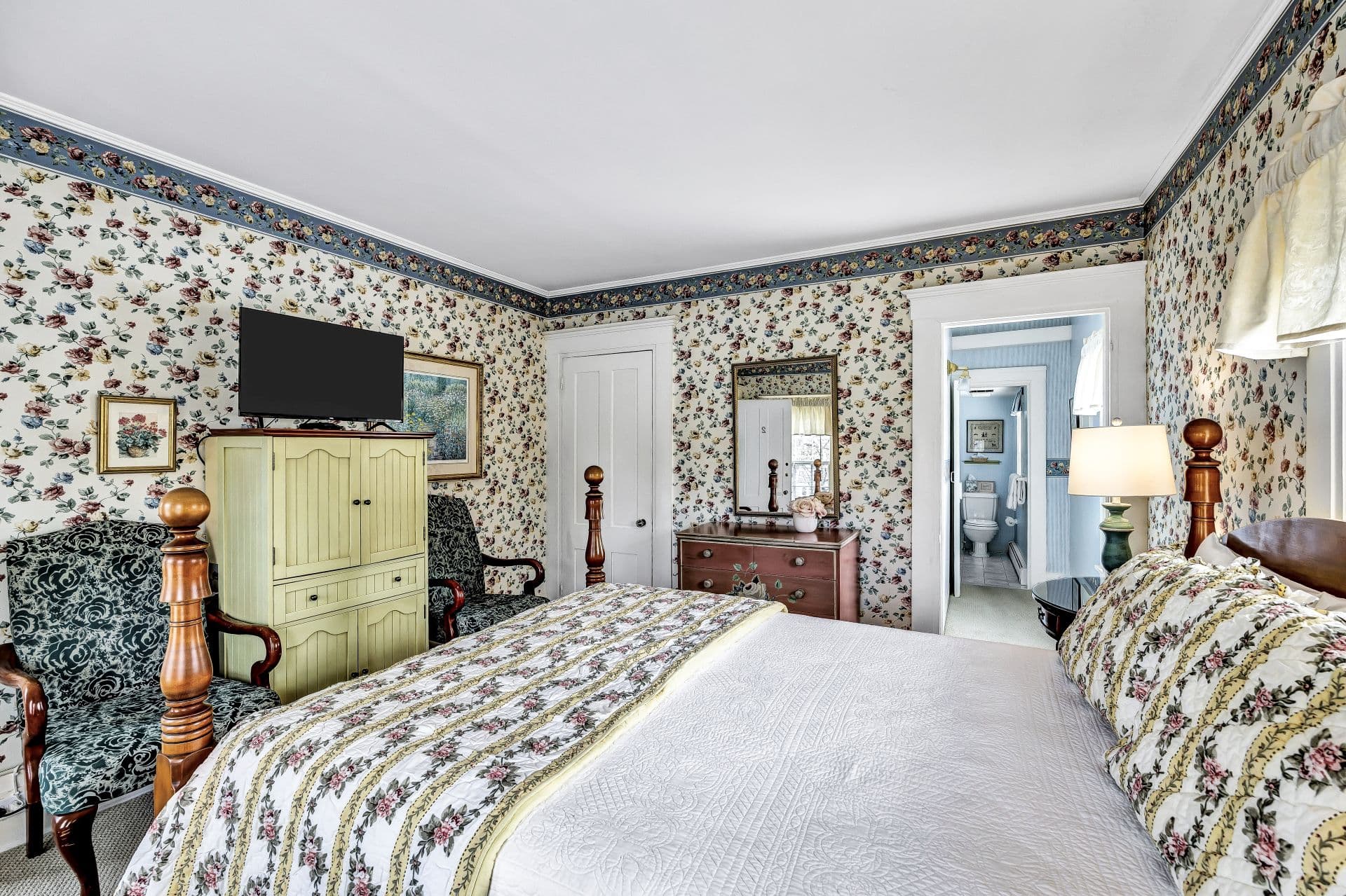 side view of Queen 4 poster bed with yellow stripes and roses bedding, white with yellow, pink and blue roses wall paper, pink shabby chic dresser with mirror above,closet, green sitting chair and flatscreen TV,