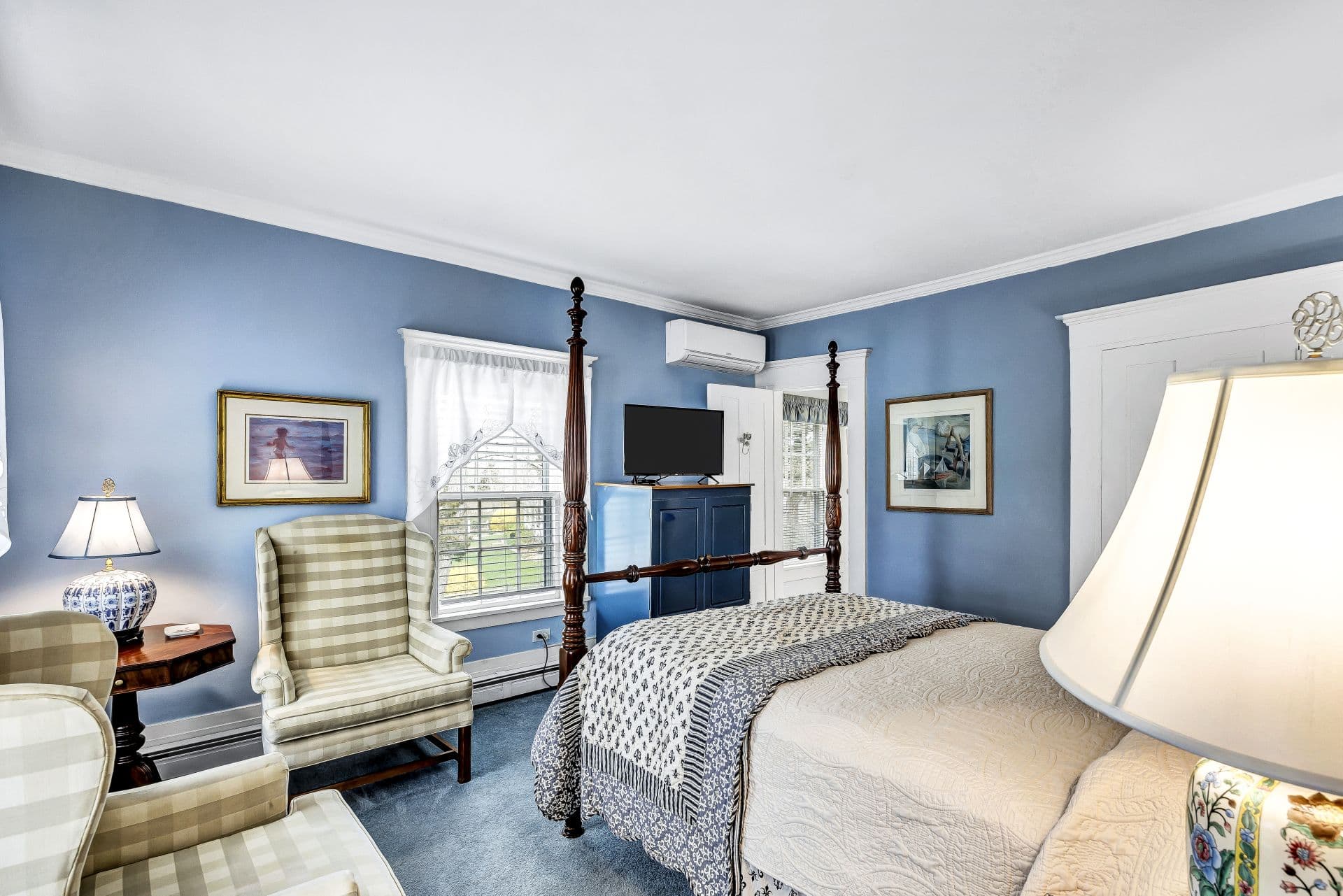 Four poster Queen bed, with blue and cream accent bedding, light blue walls and carpet, side table with lamps, flatscreen TV, reading chairs, side table with lamp,ductless A/C unit,door to bathroom