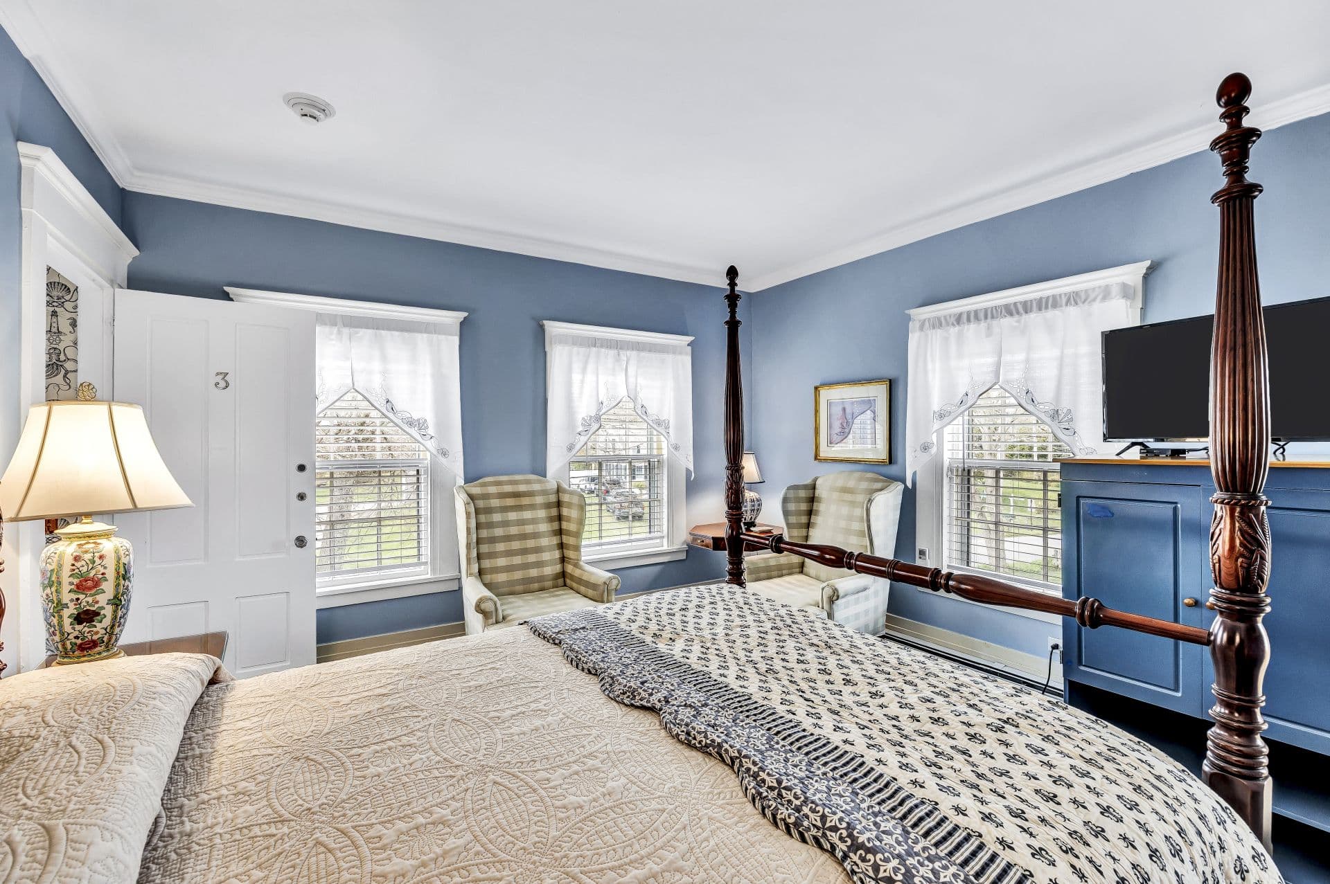 Four poster Queen bed, with blue and cream accent bedding, light blue walls and carpet, side table with lamps, reading chairs, side table with lamp, three windows, flatscreen TV