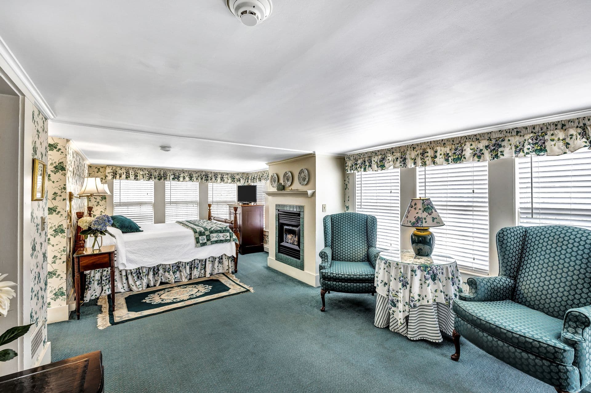 A bedroom with blue carpet and a small sitting area. The bed has a white comforter, and there's a fireplace and two blue armchairs.