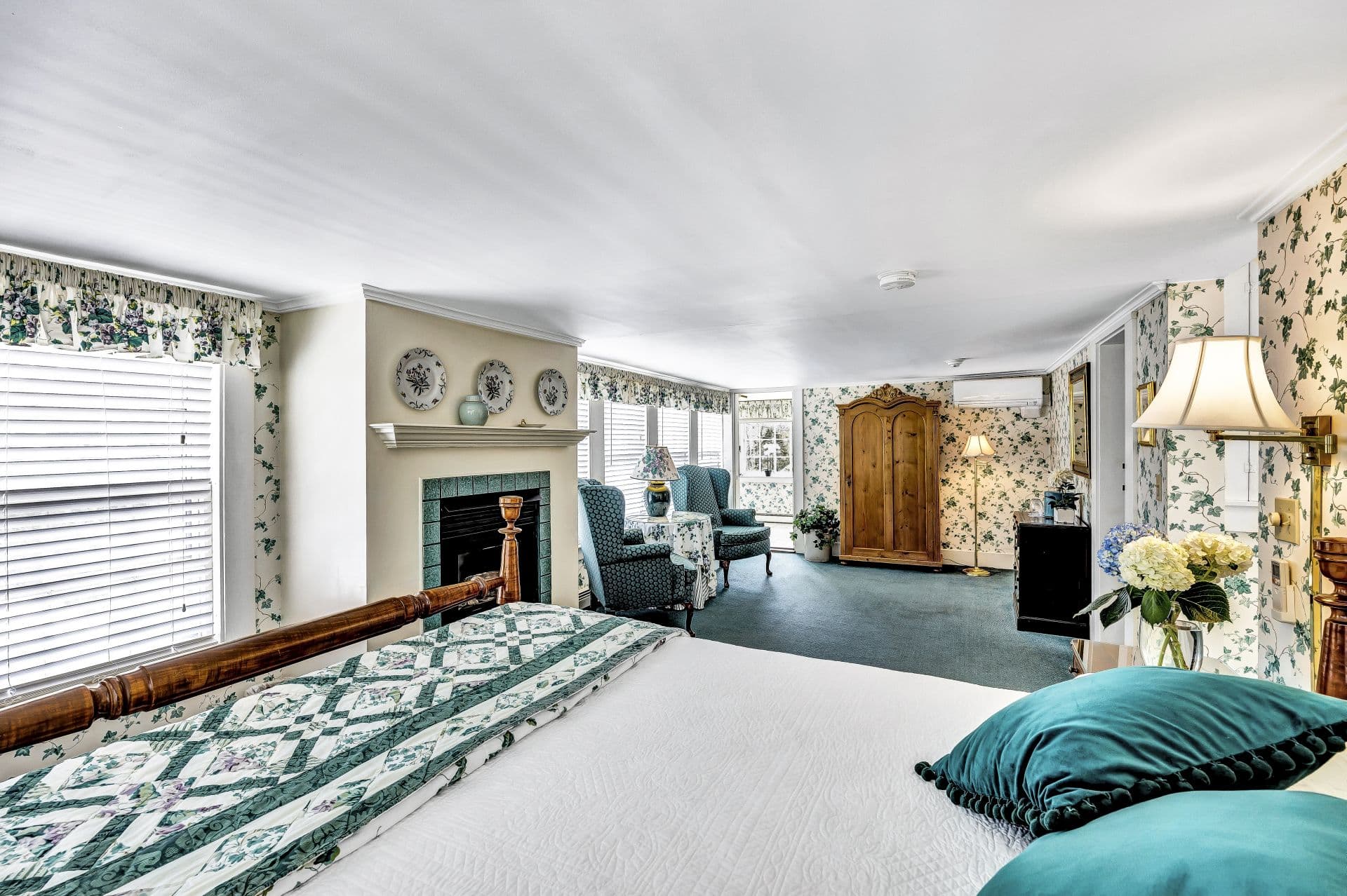 A bedroom with a fireplace and wood beams. The room has a bed with a green and white patterned blanket and a seating area.
