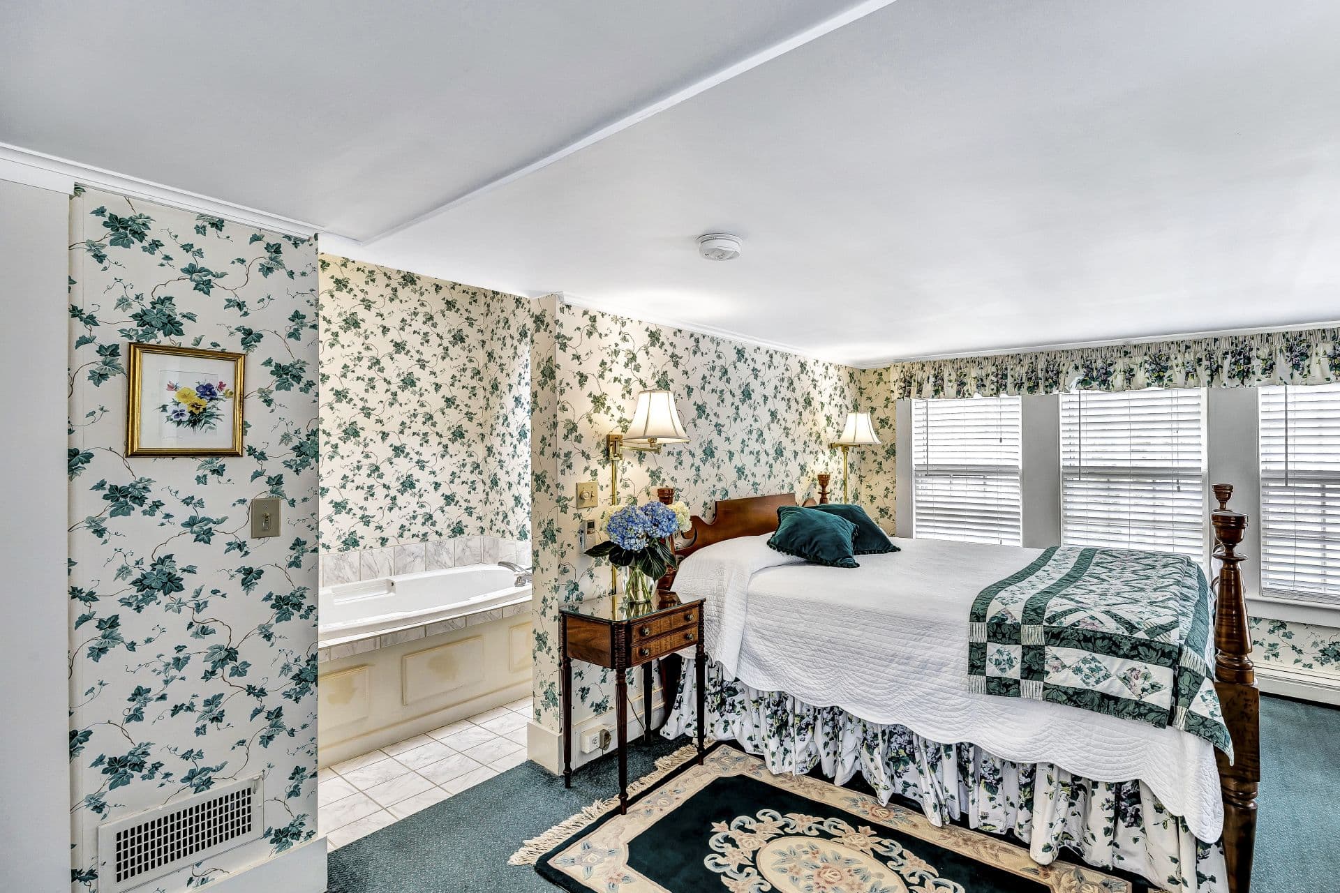 A bedroom with light green floral wallpaper, a bed with a white comforter and green quilt and a separate alcove with a soaking tub.
