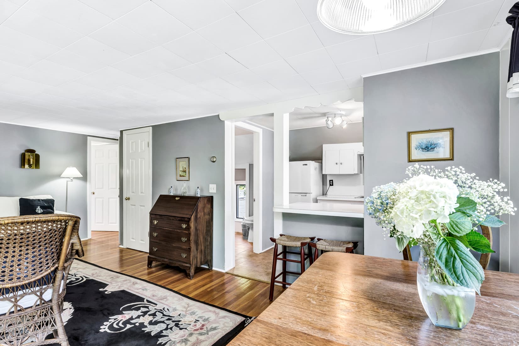 A cozy, light-filled living area featuring a wooden table, a decorative rug, and access to a kitchen in the background.