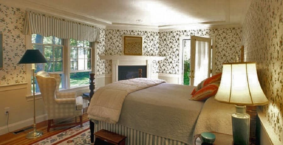 A cozy bedroom with floral wallpaper, a bed, armchair, lamp, and a view of greenery through the windows.