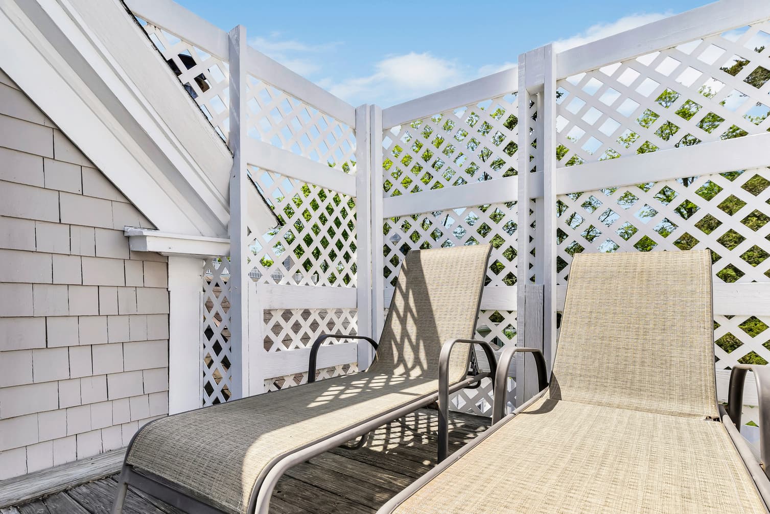 Two sun loungers on a wooden deck surrounded by a white lattice fence under a clear blue sky.