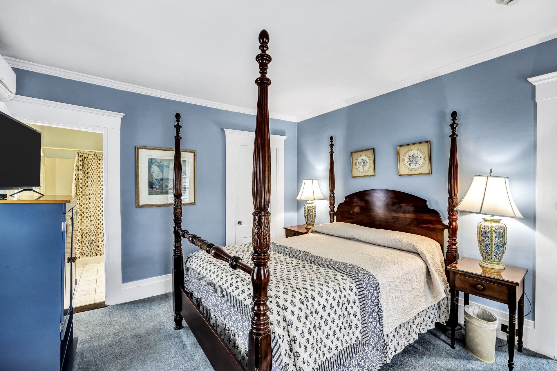 Four poster Queen bed, with blue and cream accent bedding, light blue walls and carpet, side table with lamps, door to bathroom and closet