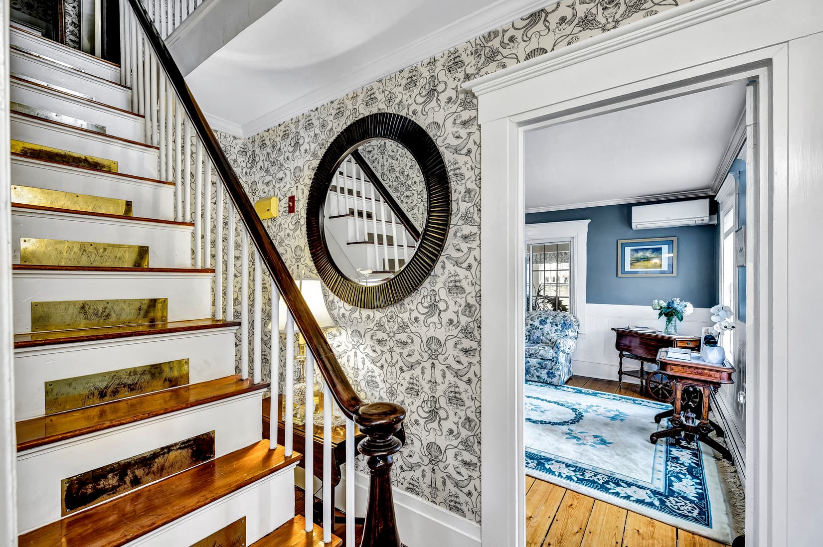 A view of a staircase leading to a mirrored wall with decorative wallpaper and an open doorway to a stylish living area.