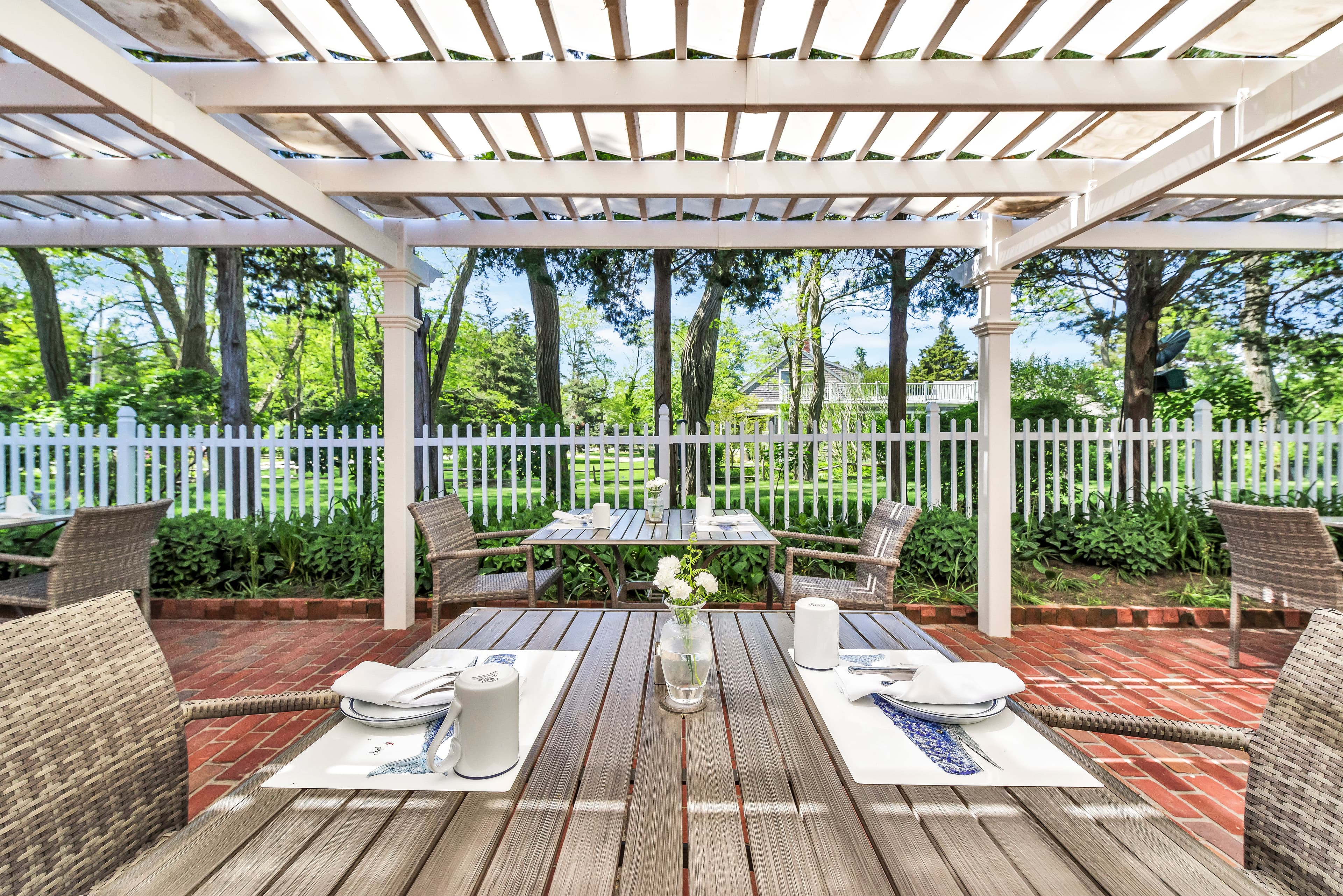 Outdoor dining area with wooden tables, surrounded by greenery and a white picket fence.