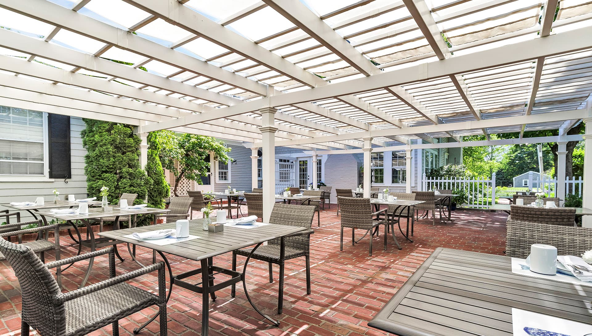A sunny patio with wicker tables and chairs, set for dining under a wooden pergola.
