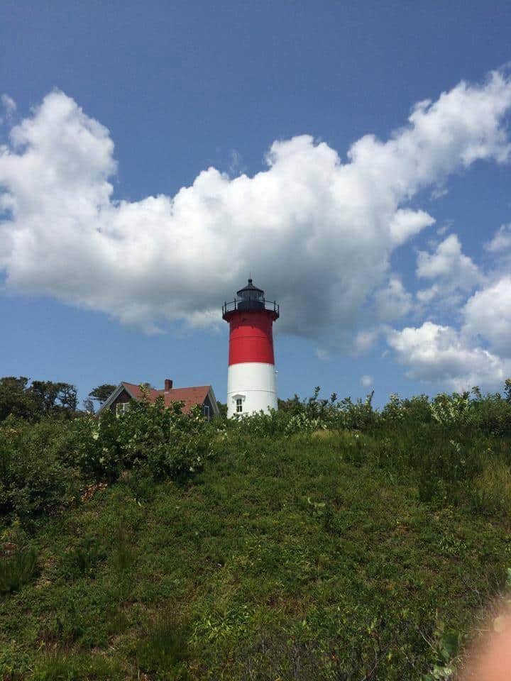A red and white lighthouse stands atop a grassy hill under a blue sky with fluffy clouds.