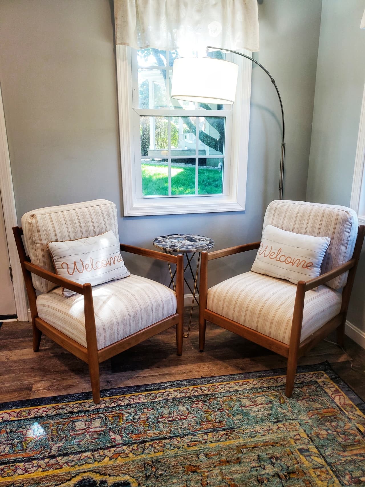 close up of sitting area with arc lamp with round shaded light, 2 pale cream stripped upholstered chairs with wooden arms and legs, blue agate small round table in center, behind is a window with pull down shades and cream valances