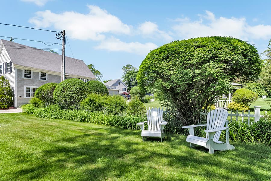 A serene yard featuring two white Adirondack chairs surrounded by green shrubs and grass with a house in the background.
