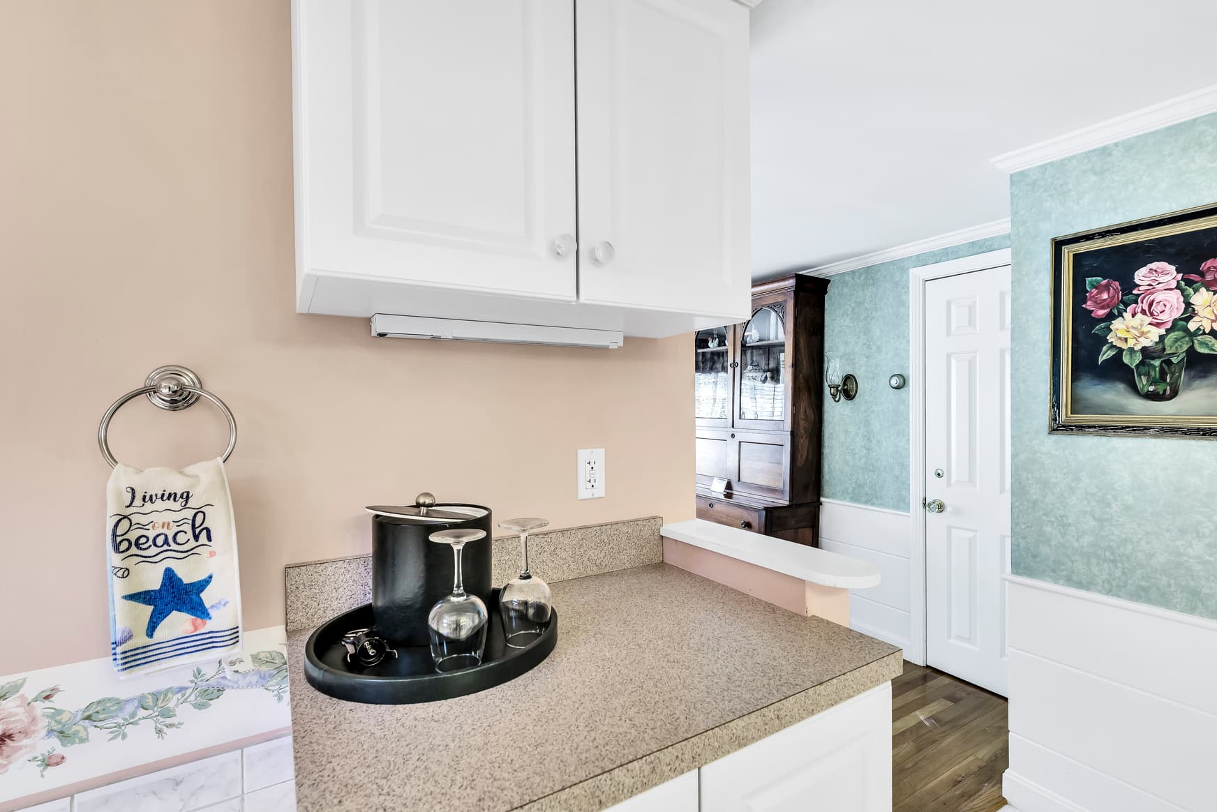 A modern kitchen corner featuring beige walls, white cabinetry, and a decorative towel with beach-themed text.