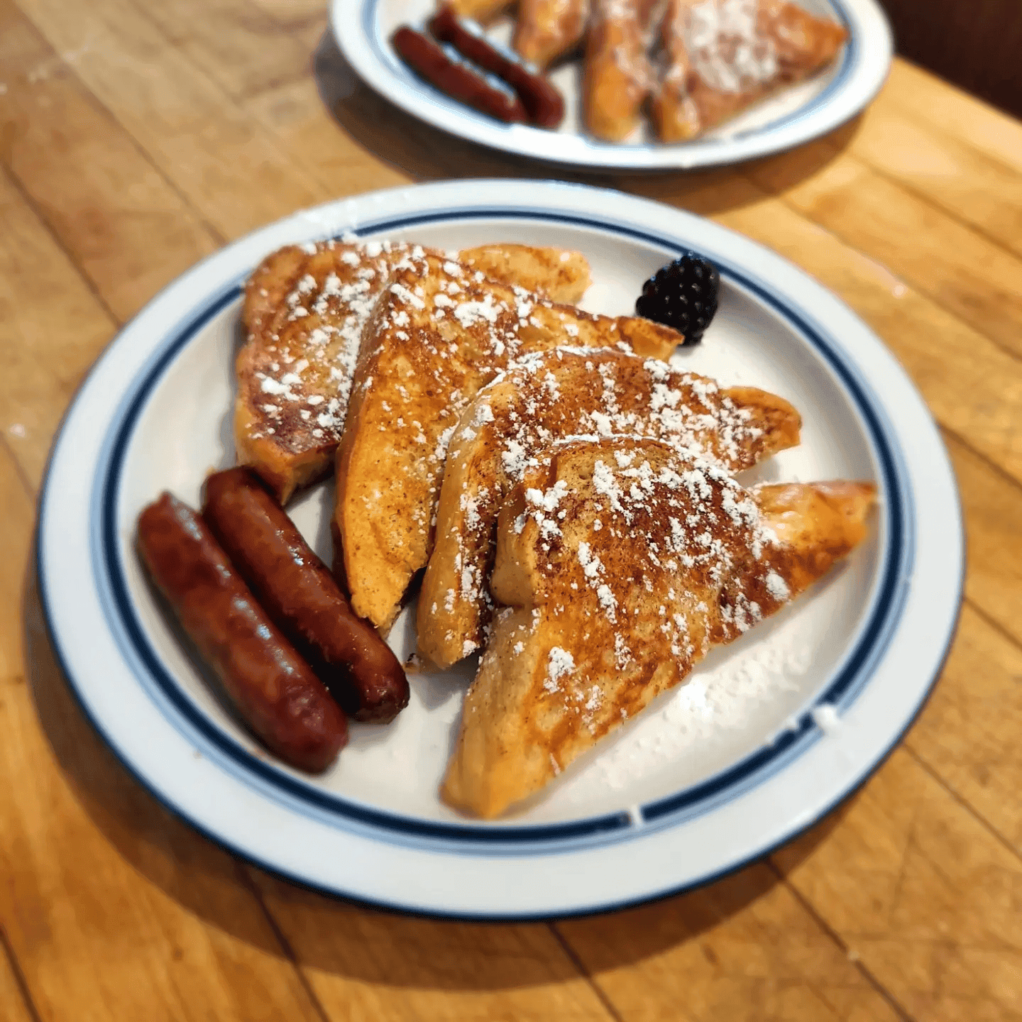A plate of French toast dusted with powdered sugar, served with sausage links and a black raspberry.